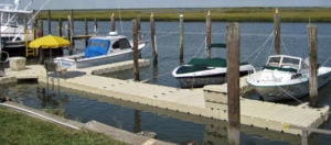  Several boats are tied up to a beige plastic floating dock, which is held in place by lines tethered to wooden pilings. 