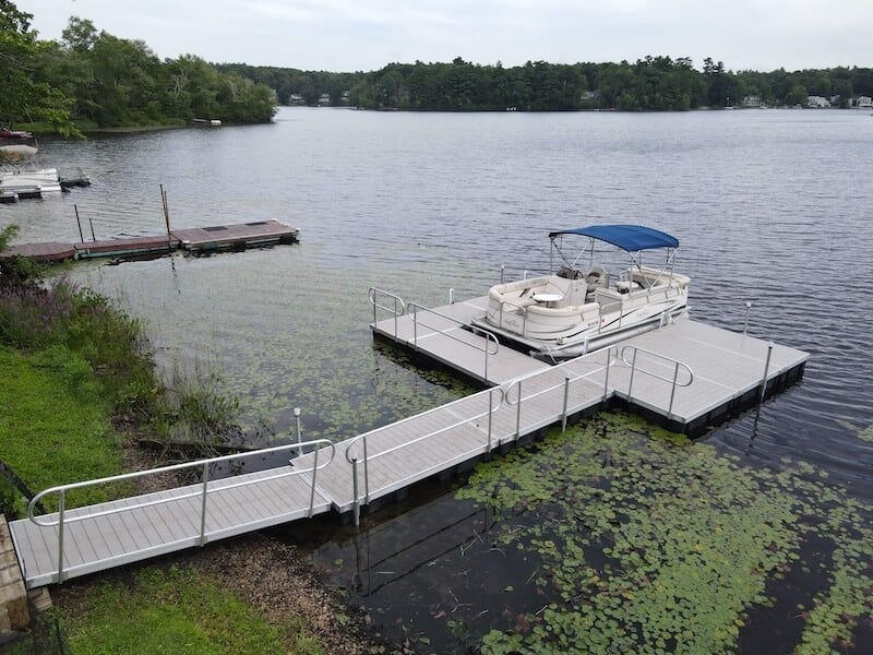 A floating dock extends from the shoreline into a lake, ending in a T-shaped platform where a pontoon boat is moored. The dock is made of light-colored material with railings for safety. The water around the dock has lily pads, and the background shows a tree-lined shore with other docks and boats in the distance. This image illustrates a floating dock, highlighting its adaptability to changing water levels and its stable platform for mooring boats.