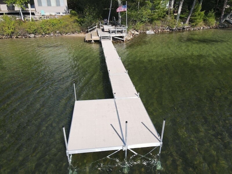 A long, narrow fixed dock extends from the shoreline into clear, shallow water, ending in a wider rectangular platform. The dock is made of light-colored material with support posts and railings along the sides. The shoreline features a small deck area with an American flag and a residential building nearby. This image demonstrates a fixed dock, highlighting its solid construction and stable connection to the shore.