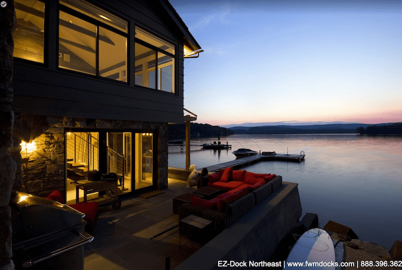A-two-story-waterfront-property-with-an-outdoor-patio-and-floating-dock-at-evening.-There-is-a-small-docked-motorboat-and-inflatable-raft-on-the-dock.