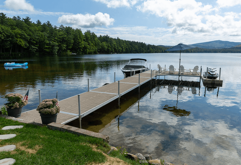 An-L-shaped-dock-on-a-lake-has-a-docked-motorboat-and-a-launch-for-a-personal-watercraft.-Four-chairs-and-an-umbrella-are-placed-at-the-end-of-the-dock-facing-the-water.