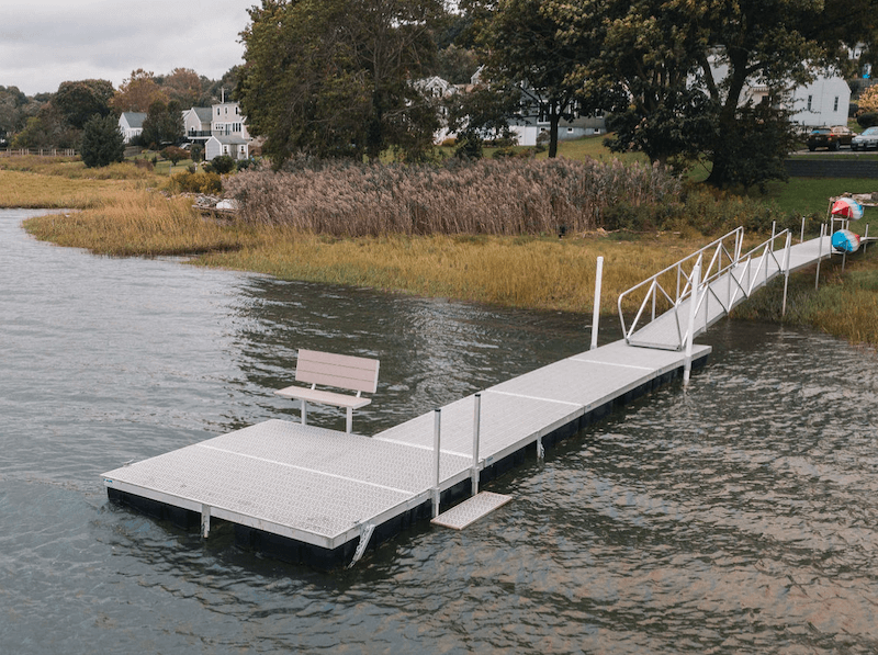 An-I-shaped-dock-on-a-lake-with-a-square-platform-placed-at-the-end-with-an-attached-bench.-A-kayak-stand-on-the-shore-holds-two-kayaks.