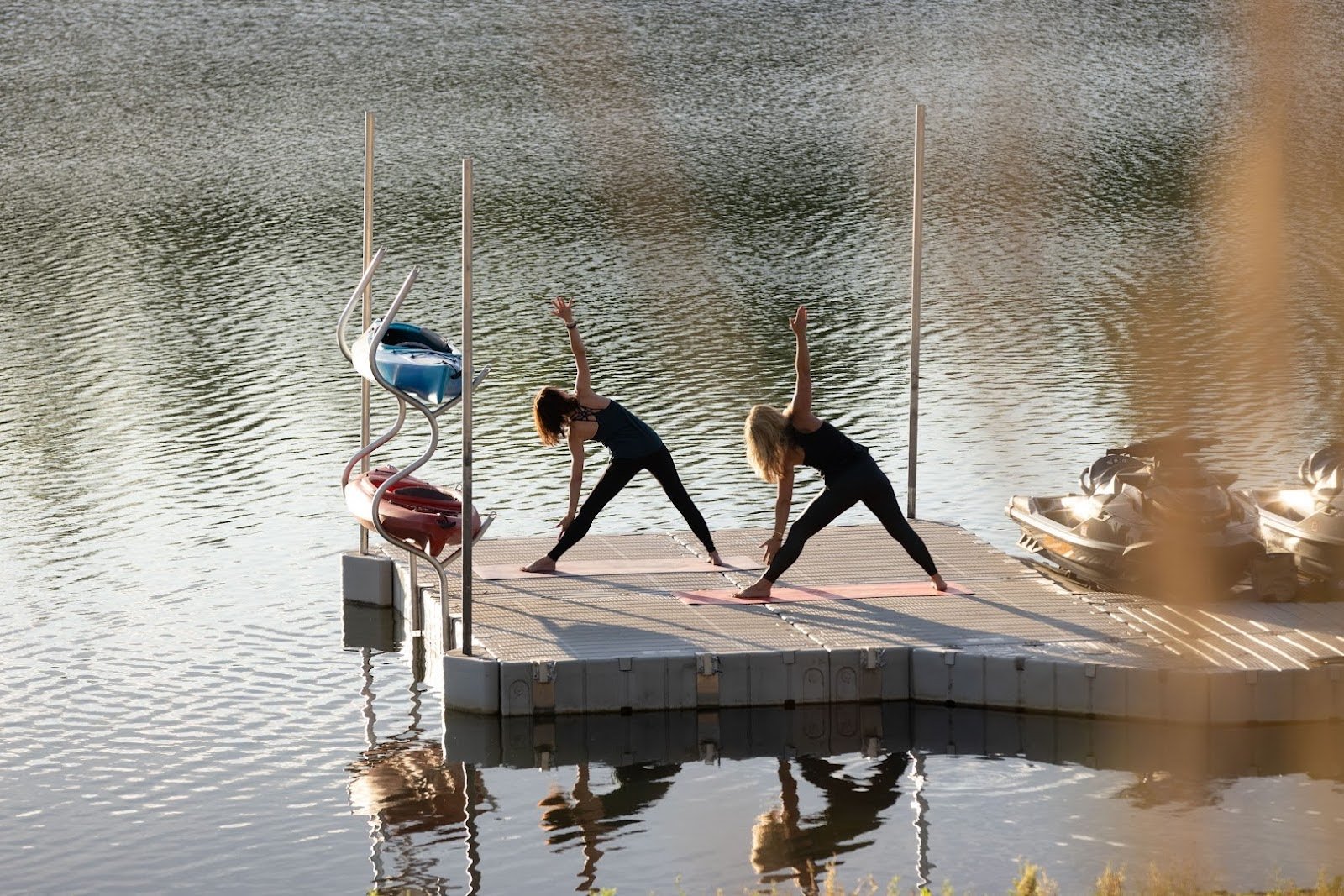 A-distant-shot-of-two-women-practicing-yoga-on-a-floating-dock.-Two-kayaks-are-resting-on-stands-and-two-jet-skis-are-moored-nearby.