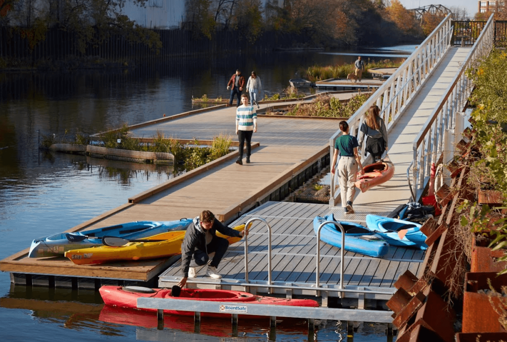 A wooden dock with wooden edge protection connects to a floating plastic EZ dock, which has an ADA-accessible ramp leading to shore. Several people walk on the wooden dock while a man docks a red kayak and two women carry an orange kayak up the ramp.